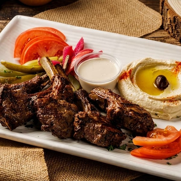 Grilled lamb chops with hummus, sliced tomatoes, pickles, and a white dipping sauce on a plate.