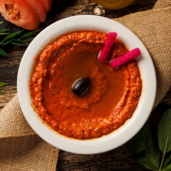 Red dip with an olive and pink pickles in a white bowl on a wooden table with burlap.