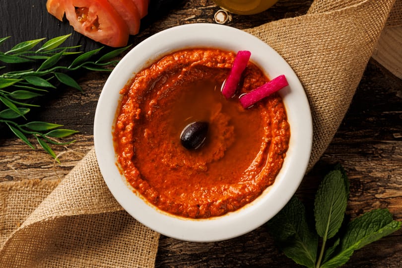 Red dip with an olive and pink pickles in a white bowl on a wooden table with burlap.