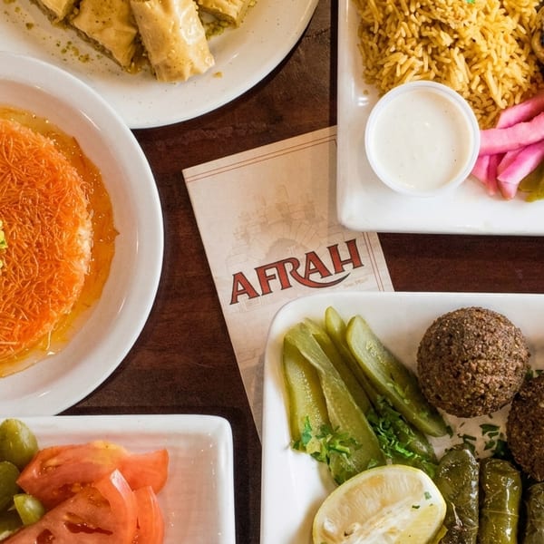 Afrah restaurant menu surrounded by plates of falafel, kebabs, rice, pickles, and kunafa dessert.