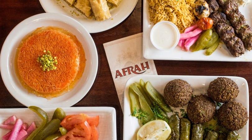 Afrah restaurant menu surrounded by plates of falafel, kebabs, rice, pickles, and kunafa dessert.