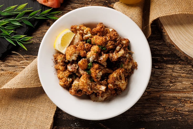 Seasoned roasted cauliflower with a lemon wedge on a white plate, set on a rustic wooden table.