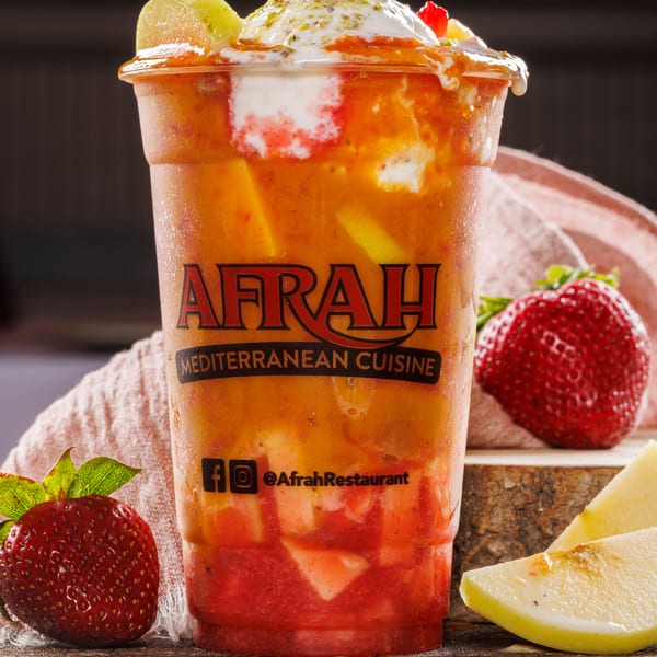 Afrah Mediterranean Cuisine layered fruit drink topped with cream, pistachios, and kiwi, beside strawberries and apple.