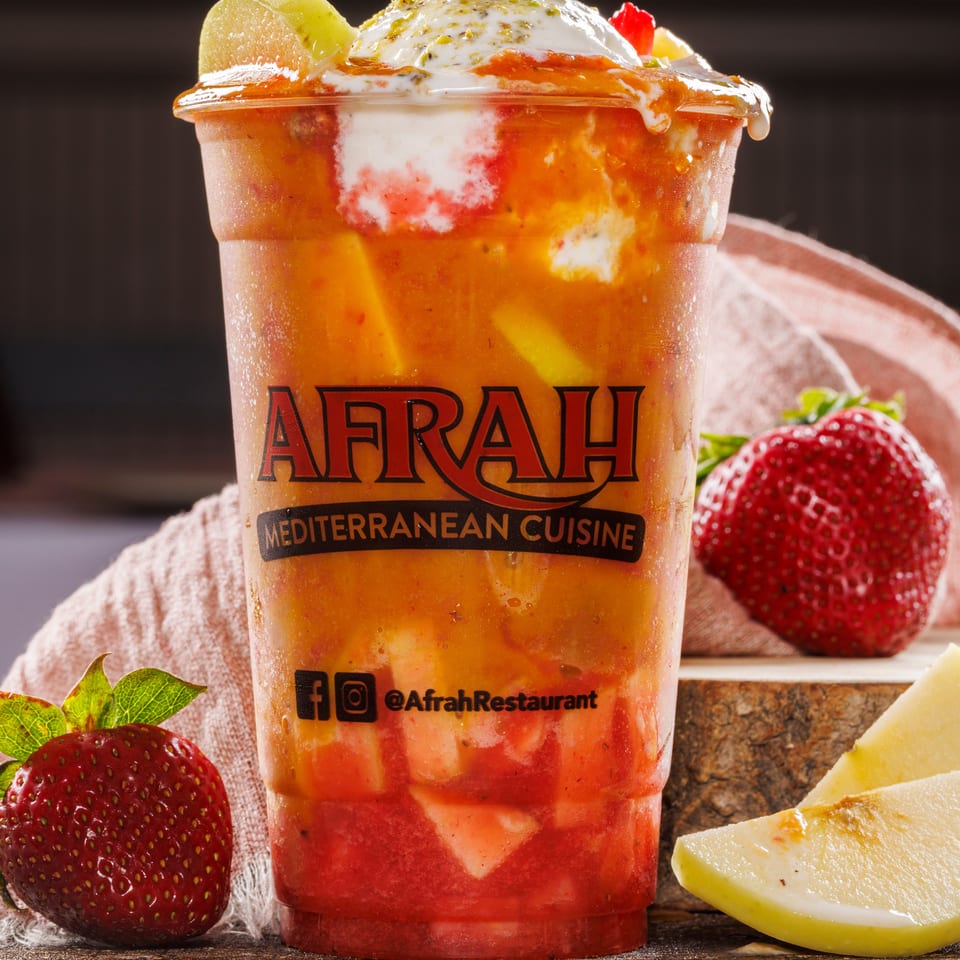 Afrah Mediterranean Cuisine layered fruit drink topped with cream, pistachios, and kiwi, beside strawberries and apple.