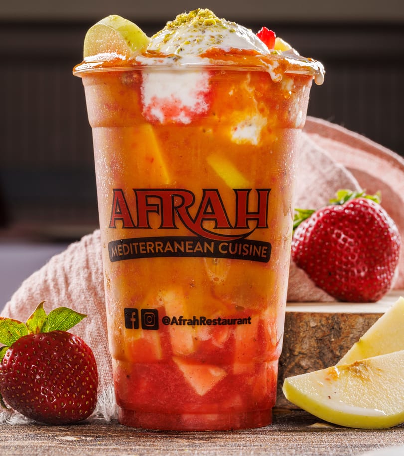Afrah Mediterranean Cuisine layered fruit drink topped with cream, pistachios, and kiwi, beside strawberries and apple.
