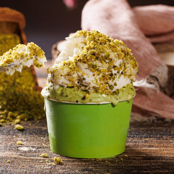 White ice cream with pistachio topping in a green cup, a spoon lifts a scoop, with more pistachios nearby.
