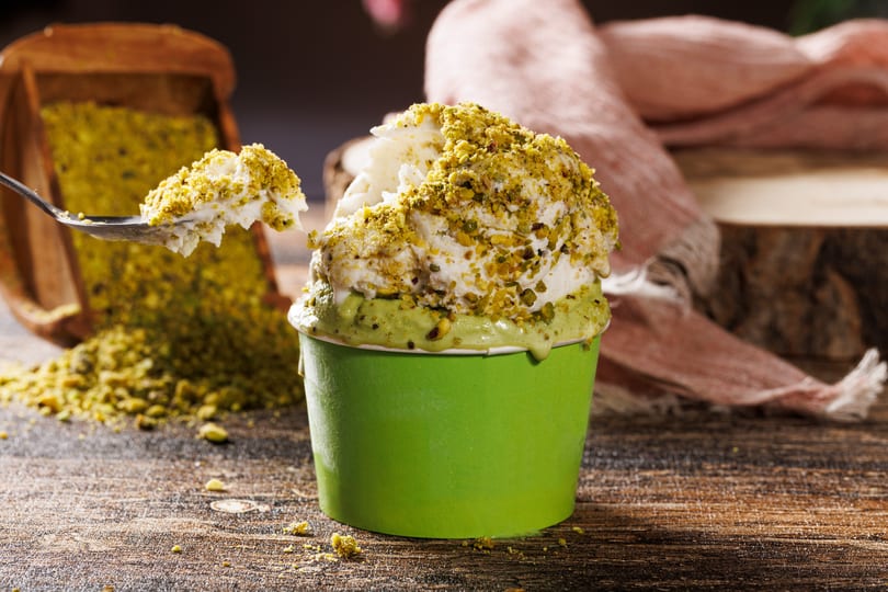 White ice cream with pistachio topping in a green cup, a spoon lifts a scoop, with more pistachios nearby.