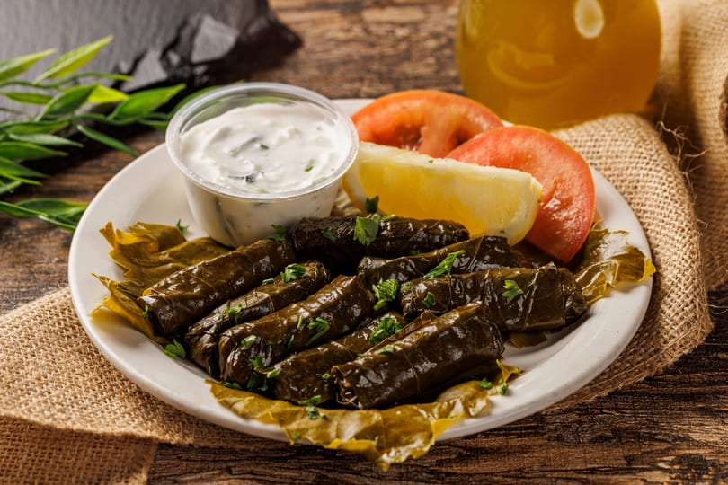 Plate of dolmades with tzatziki, lemon, and tomato slices on a rustic wooden table.