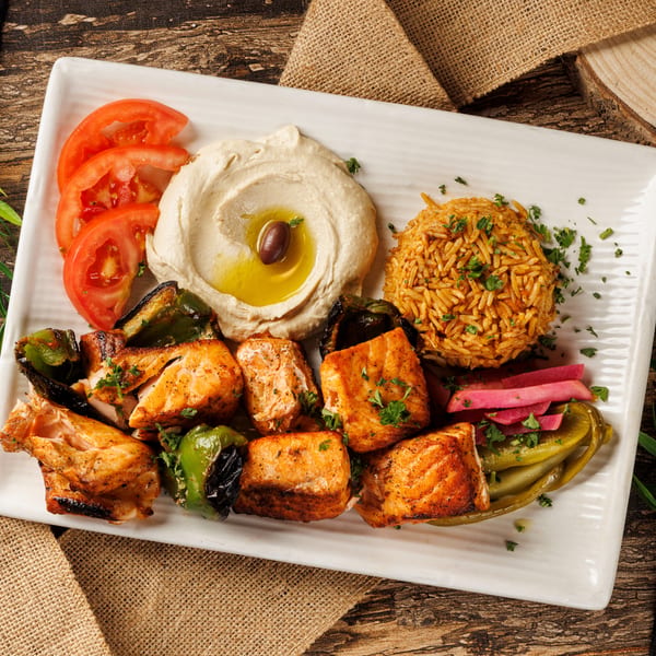 Grilled salmon skewers, rice, hummus, tomatoes, and pickles on a white rectangular plate.