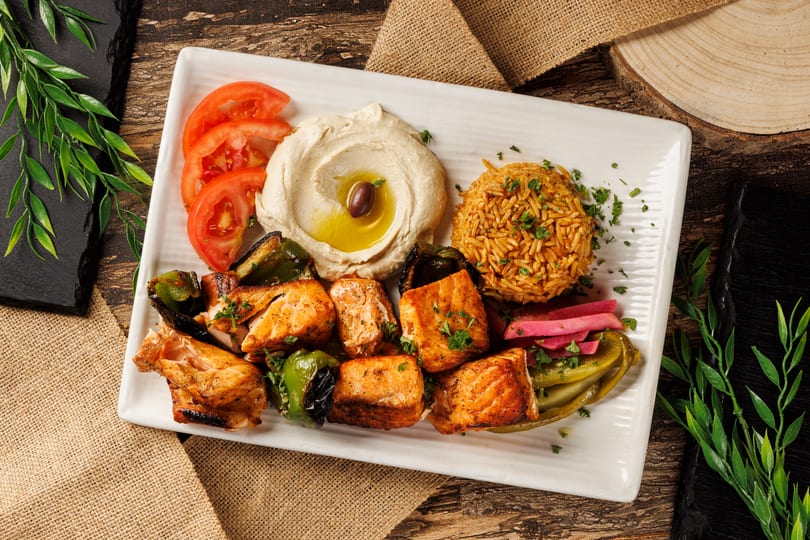 Grilled salmon skewers, rice, hummus, tomatoes, and pickles on a white rectangular plate.