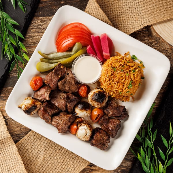 Grilled beef skewers with roasted vegetables, rice, pickles, tomatoes, and sauce on a white plate.