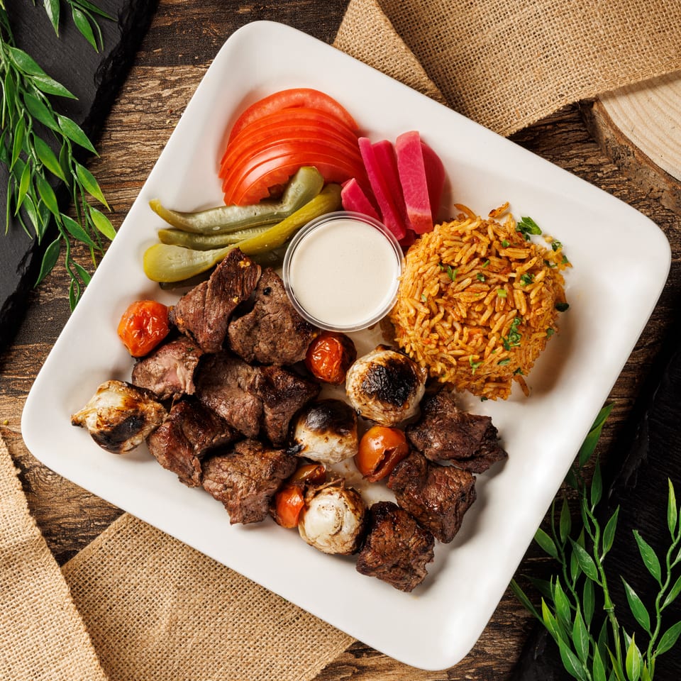 Grilled beef skewers with roasted vegetables, rice, pickles, tomatoes, and sauce on a white plate.