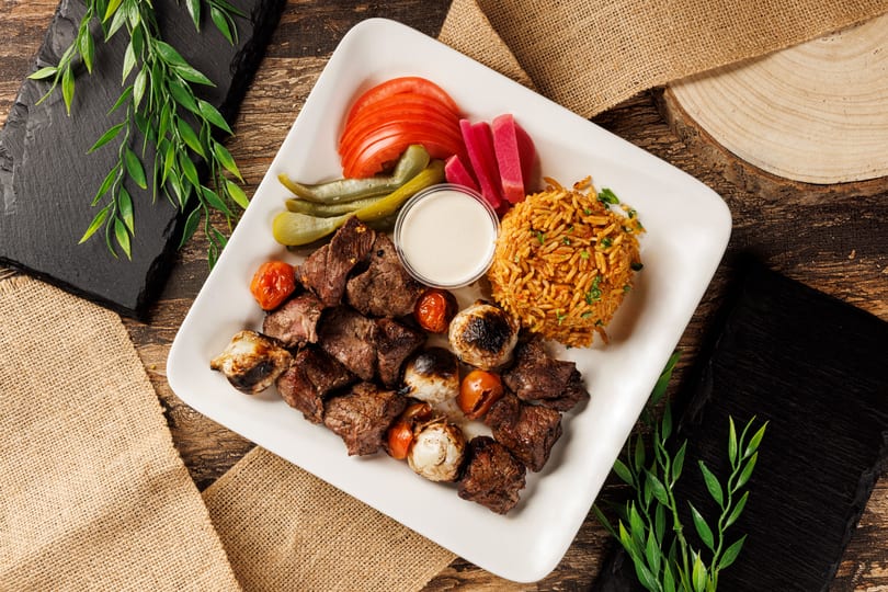 Grilled beef skewers with roasted vegetables, rice, pickles, tomatoes, and sauce on a white plate.